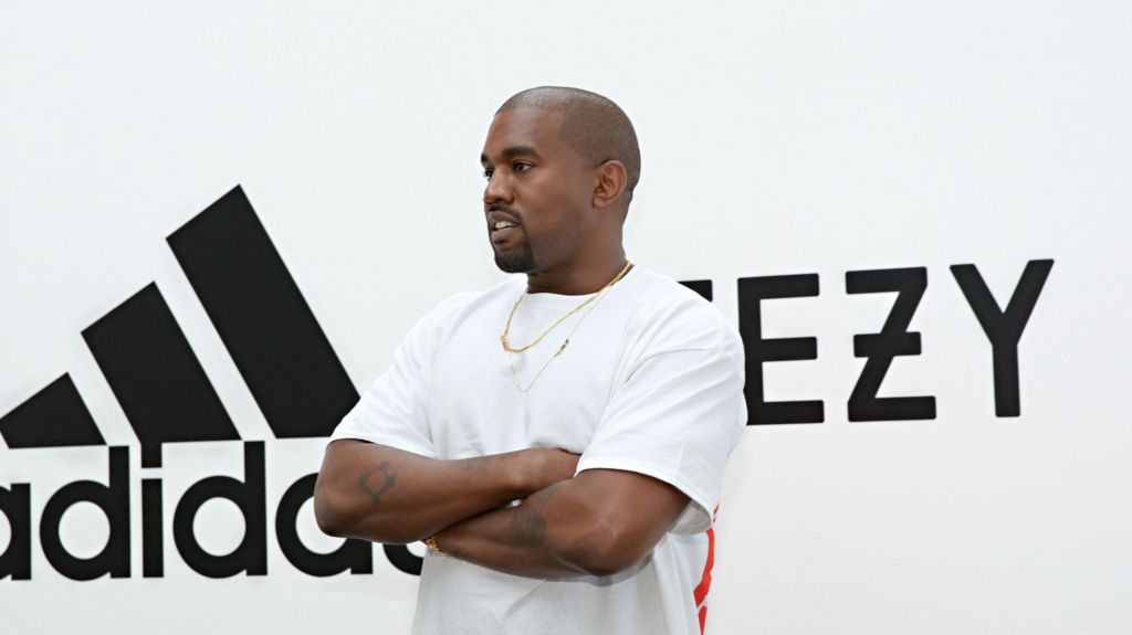 West in a white T-shirt with arms folded in front of Adidas/Yeezy sign