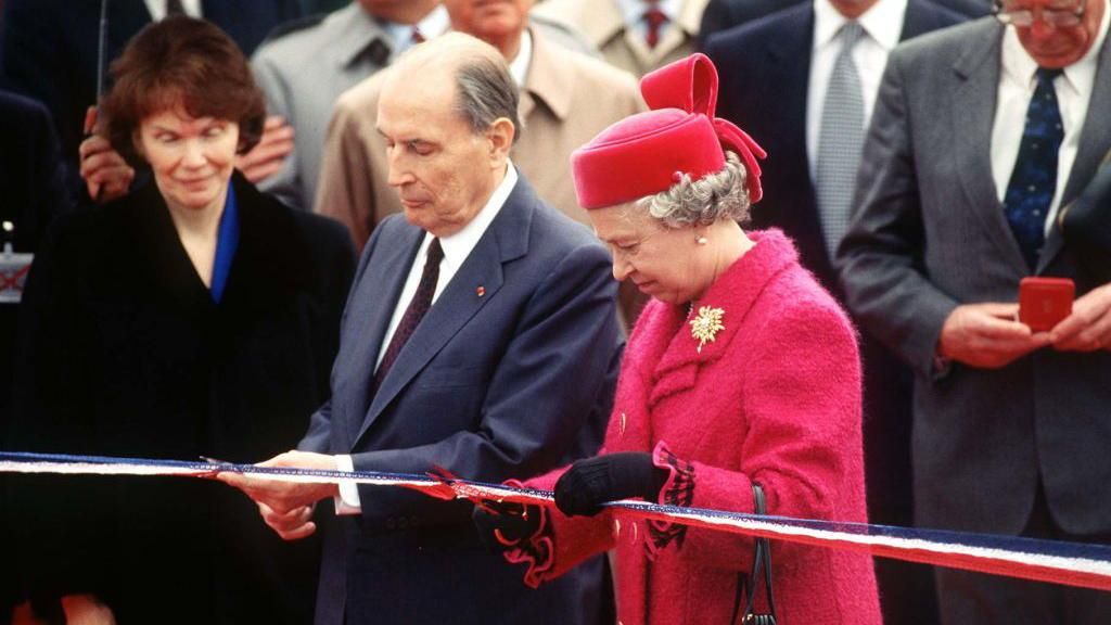 President Mitterrand in a blue suit and burgundy tie stands beside Queen Elizabeth in a red dress coat and hat as they cut a ceremonial ribbon at the opening of the Channel Tunnel