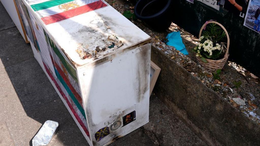 A cabinet with images of the Iranian flag attached is covered in dark charred markings. Flowers can be seen lining a small ledge.