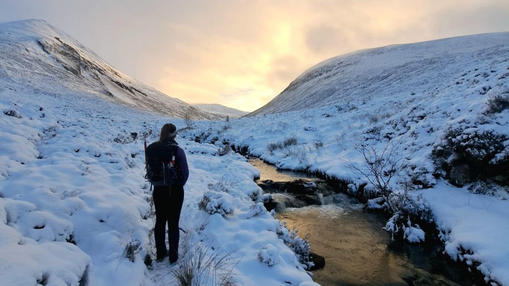 A woman stands with her back to the camera in a snowy landscape. a river runs down past her left hand side. In front of her is a dramatic mountain pass.