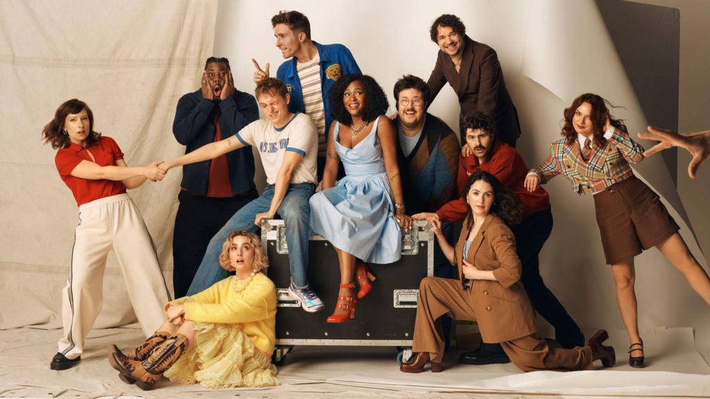 Picture of SNL UK cast set against a beige backdrop with cast members stood against a black box