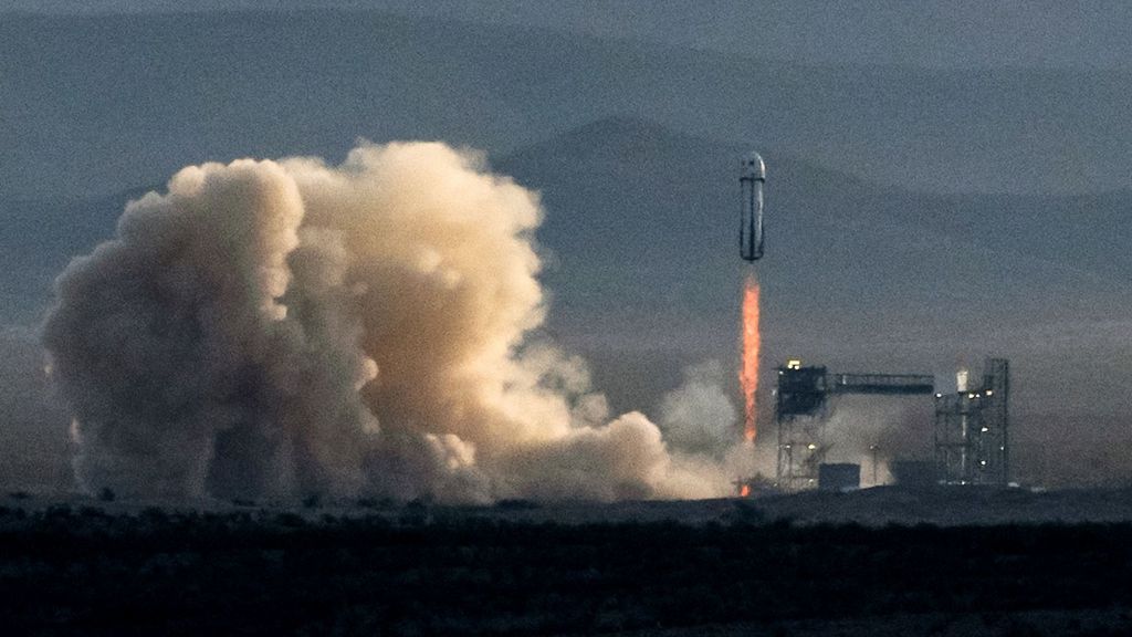 A wide view shows the rocket lifting off from the ground, with fire coming out of its bottom end and smoke pluming around it.