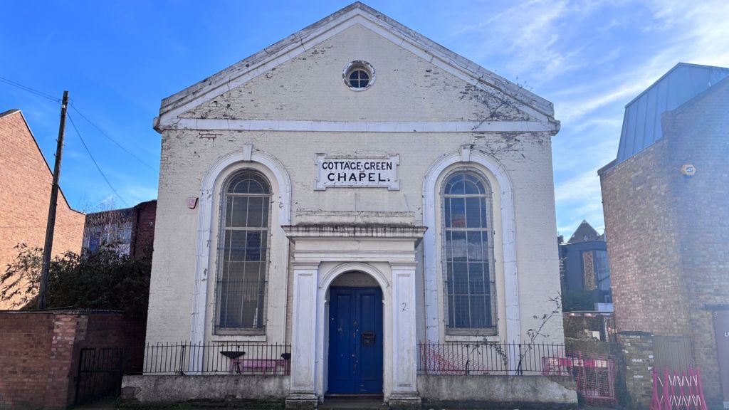 A cream coloured building with a blue door showing years of wear and tear with peeling paint and weeds growing from the cracks