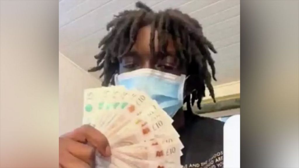 Daejaun, wearing a blue surgical mask, holds a fanned stack of £10 notes. He has medium‑length dreadlocks and is indoors, with a white ceiling and light-coloured walls in the background.