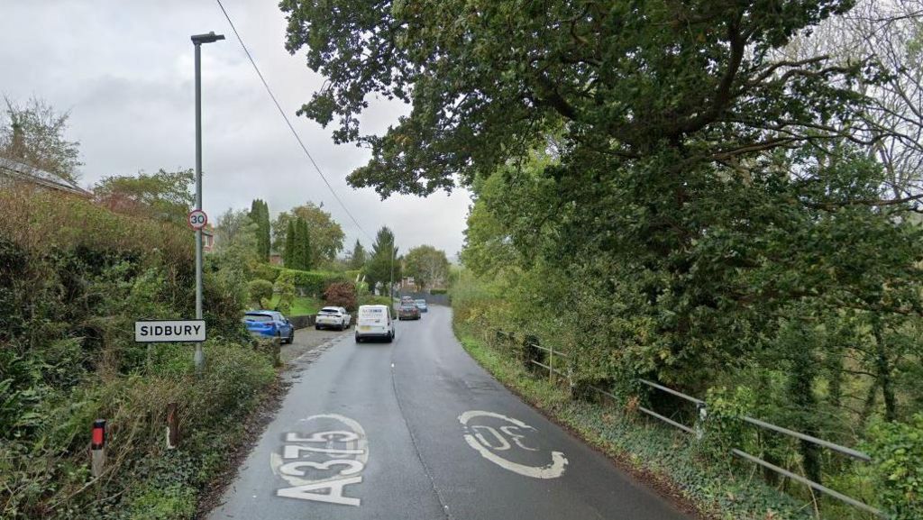 A Google street view image of the A375 in Sidbury with a road sign for the village on the left and six vehicles on the road heading away from the camera. Trees and shrubs are on both sides of the road.