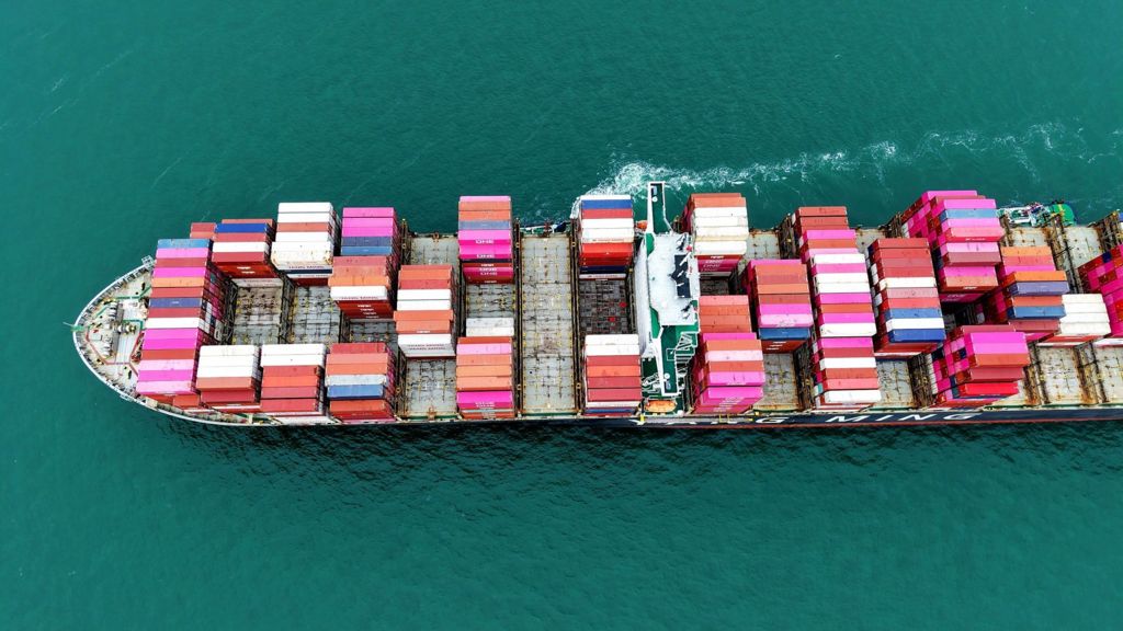 A large cargo ship staked with pink, blue and white container ships sails from right to left through the picture, a small trail of white foam in the picture.