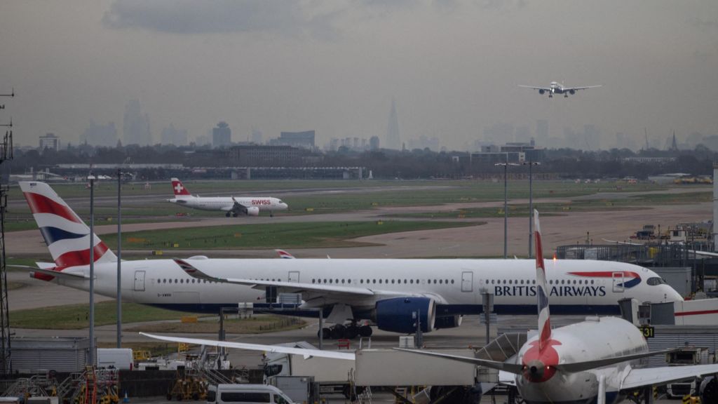 Planes arriving and departing from the runway at London Heathrow. One plane is coming into land, whilst others traverse the runways on the ground, where a BA plane parks at a gate whilst another smaller Swiss Airline plane also taxis behind it. The London skyline is clearly visible in the distance on a grey cloudy day, with the Shard being especially prominent.