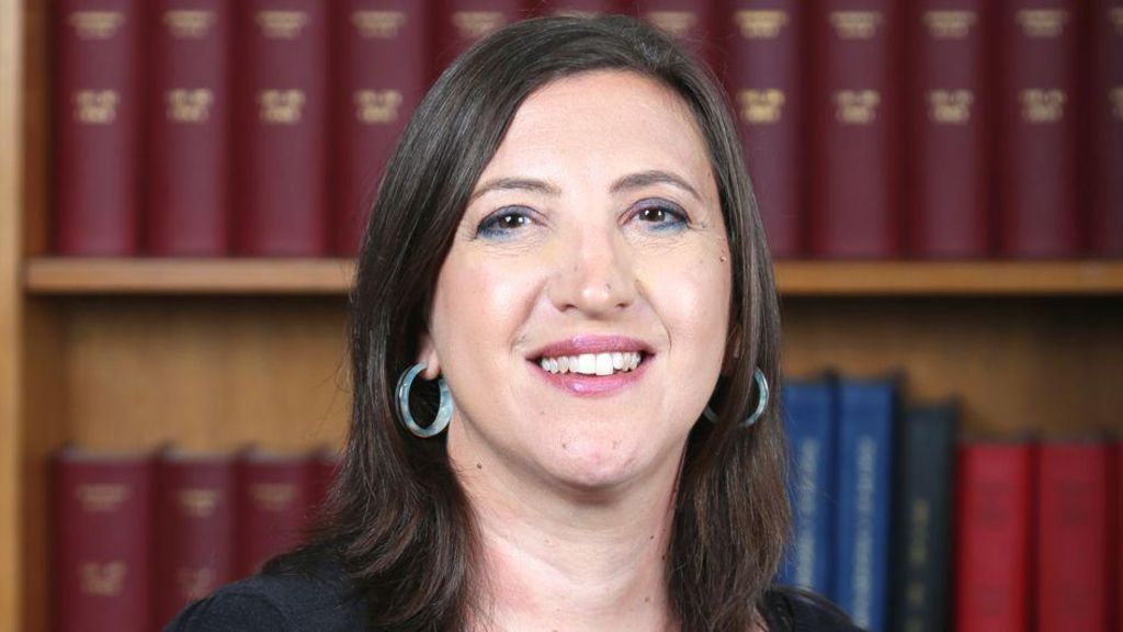 A woman in a black top and silver jewellery smiles at the camera. She has a bookcase behind her. 