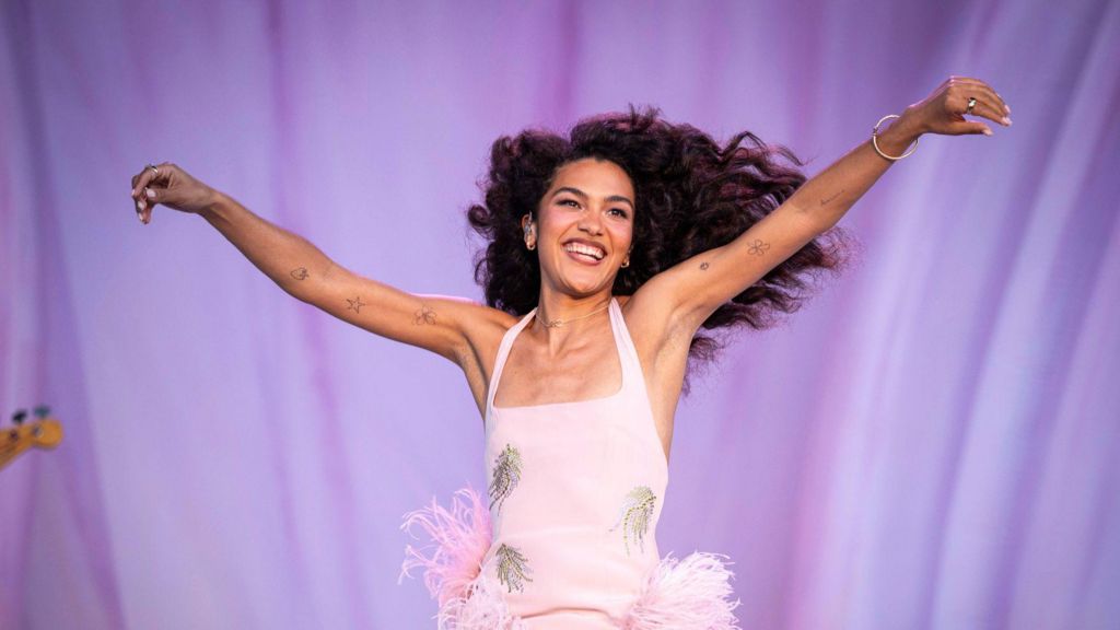Olivia Dean stands with her arms wide, smiling. Her long dark hair is blowing to the side. She's wearing a pink dress.