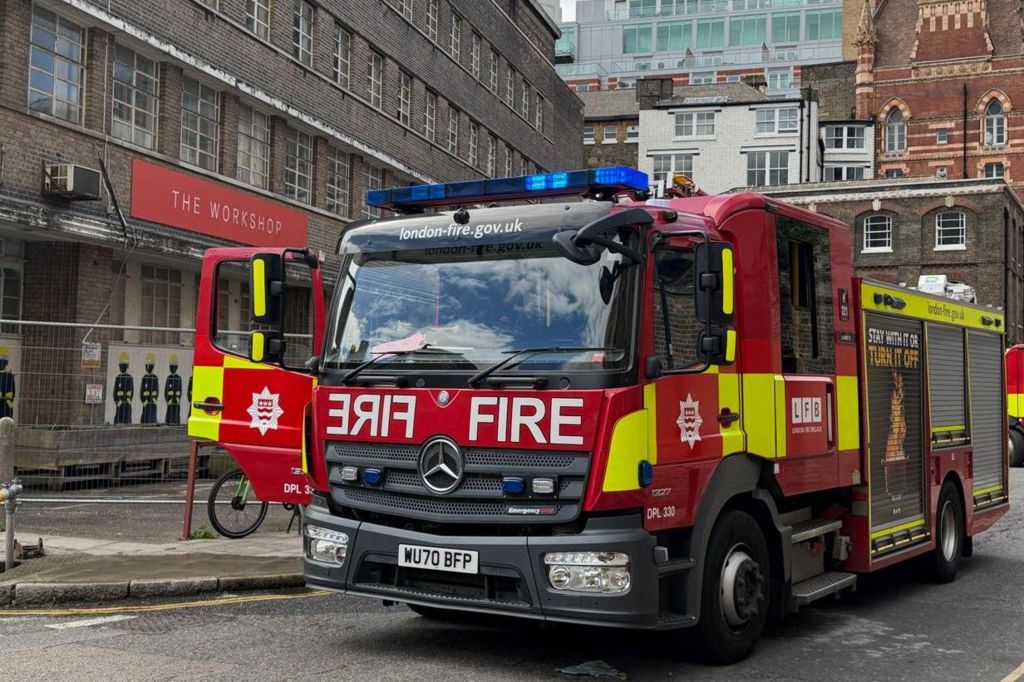 An LFB fire engine parked on a road in London with buildings in the background. The driver side door is open.