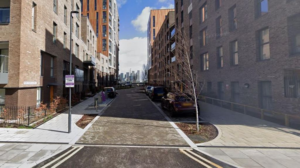 New Union Close which shows high rise apartments on both sides, a paved road and cars parked in bays. In the far distance there is the London skyline of tower blocks and blue sky.