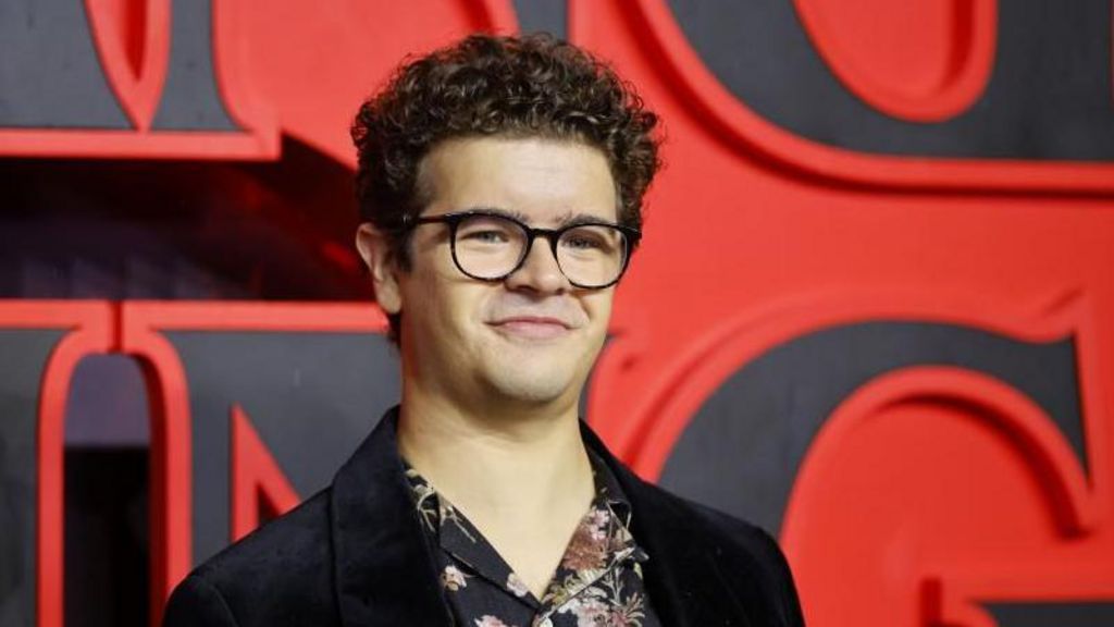 Gaten Matarazzo has short curly hair and is wearing black-rimmed glasses, a black Velvet blazer and floral shirt underneath. He is standing on a red carpet in front of a huge 'Stranger Things' sign
