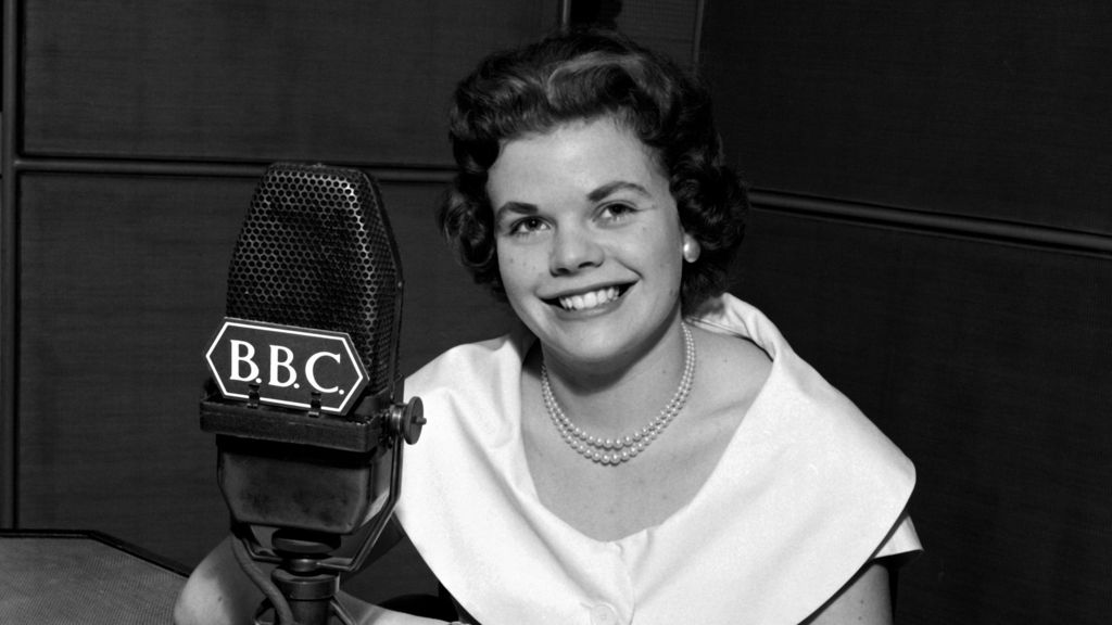 Black & white photo of a young Judith Chalmers in front of a BBC microphone in 1957