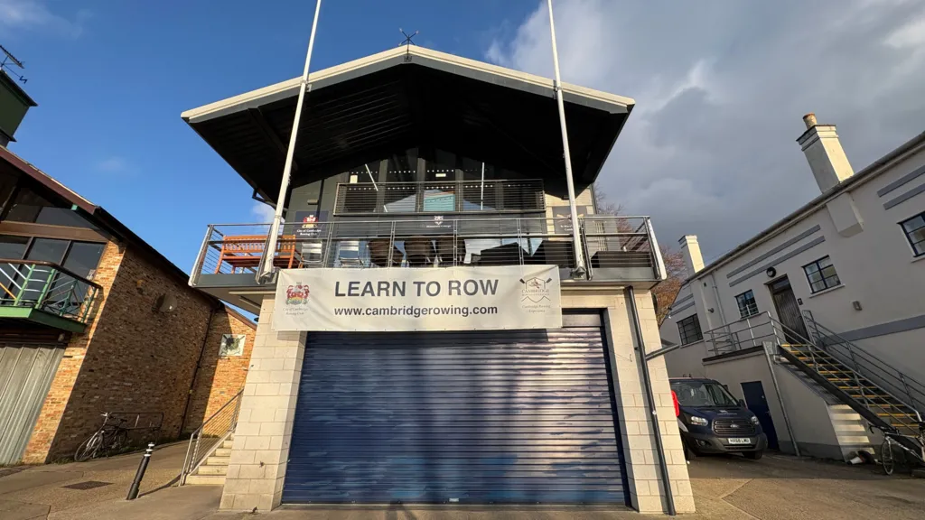 A building, centre frame and shot from below. It is a boathouse on the River Cam, though the water is not in frame. It has a blue, roller shutter type door, over which a banner reading 