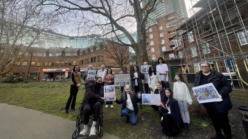 A group of adults and children, one in a wheelchair, hold placards on  grassy verge in the middle of a council estate.