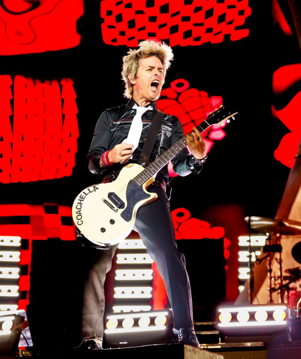Billie Joe Armstrong of Green Day pulls a face while strumming a white electric guitar.
