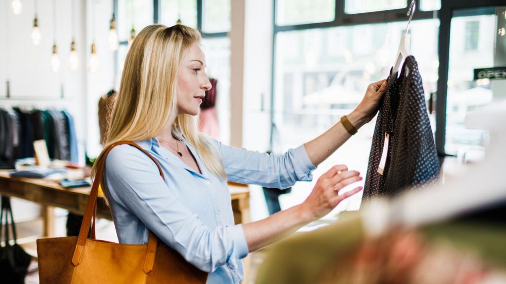 A woman with long blond hair looks at a top in a clothes shop. The woman is wearing a light blue top and has a light brown bag on her shoulder