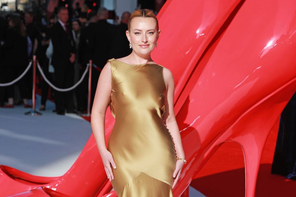Amelia Dimoldenberg poses on the red carpet wearing a gold, satin dress.