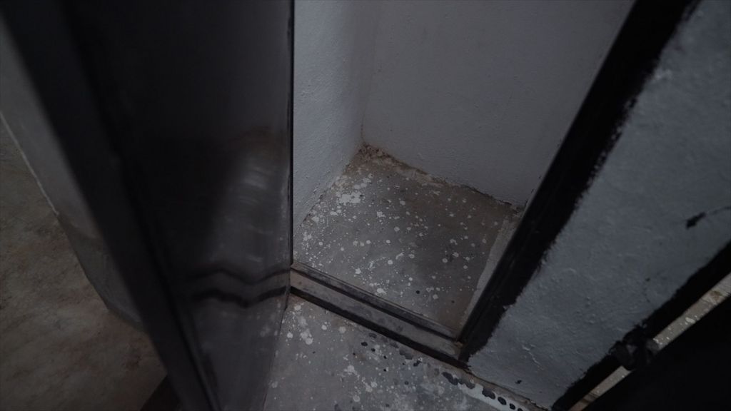 Photo looking down showing the lower part of a cell that is about a metre by a metre in size, with a concrete floor and white-painted walls. Its tall black door is open.