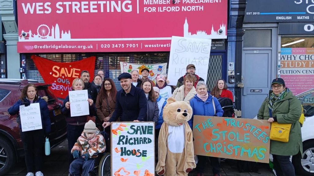Protesters stand outside Wes Streeting’s constituency office holding banners reading “Save Richard House” and “Save our services”, with adults and children gathered.
