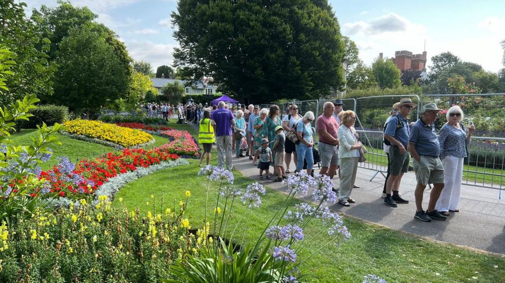 A queue of people lining up to get into the Taunton Flower Show. It's a sunny day and there are colourful displays of flowers lining the path