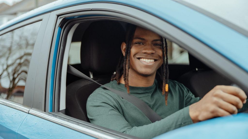A young man sitting in the driver's seat of a car. He has dreadlocks and is wearing a long-sleeve green top