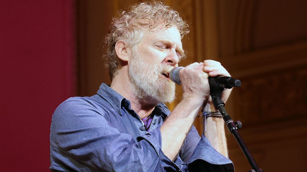Glen Hansard has short curly reddish/brown/grey hair and a greyr beard and is singing with two hands on the microphone and his eyes closed