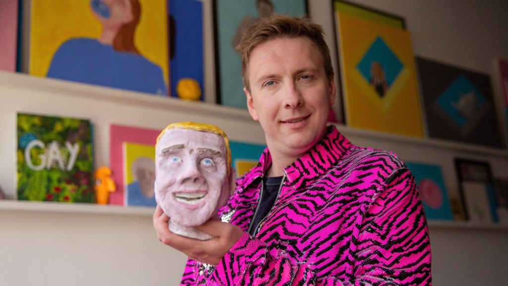 Joe Lycett wearing a pink striped jacket and holding a clay mask of his own face.