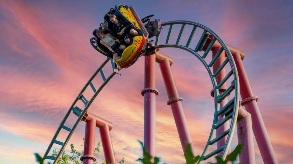 An image shows two riders inside a car on the Dragon's Fury ride as it speeds around a tight bend in a track. The car is almost at 90 degrees, with the riders facing the ground and smiling. The scene is illuminated by a dramatic red sunset.
