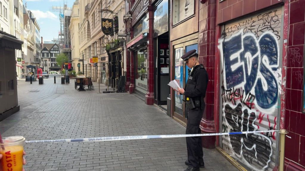 A police officer stands behind police tape on a city street in central London, reading notes outside a closed shop with graffiti on its shutter, while nearby streets are quiet and cordoned off.