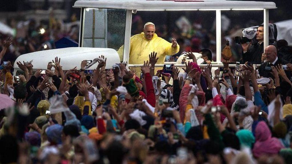 Pope Francis is seen wearing a yellow plastic rainproof in an open car. He is waving to the crowd which surrounds him.