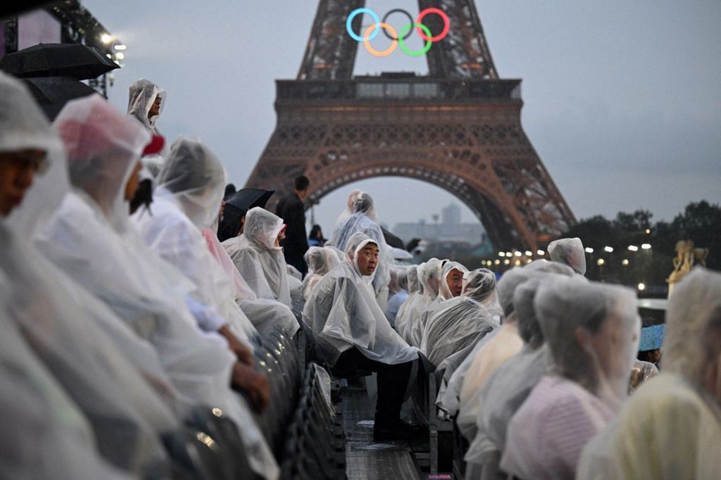 Attendees wear rain covers as they sit in the stands in front of the Eiffel Tower