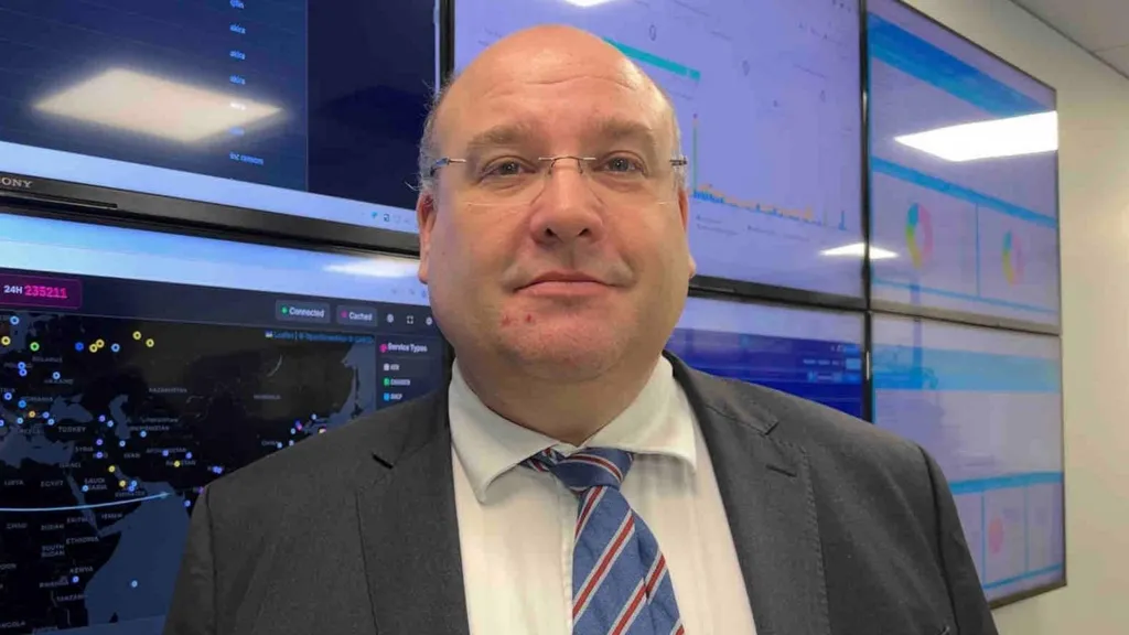 A man standing in front of multiple TVs with different graphs and numbers on them, he is wearing a suit and tie and a pair of glasses.