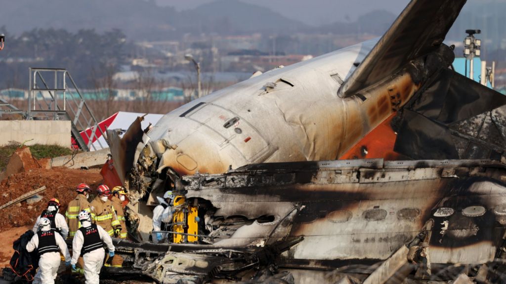 Firefighters and rescue workers in suits and helmets approach the charred wreckage of the plane.