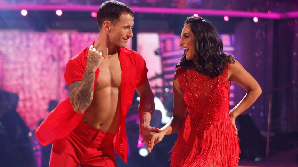 Dr Punam Krishan - wearing a red sparkly sleeveless dress with fringing, and Gorka Marquez - wearing an unbuttoned red short and red trousers - look at each other smiling as they dance in front of a purple backdrop on Strictly Come Dancing 