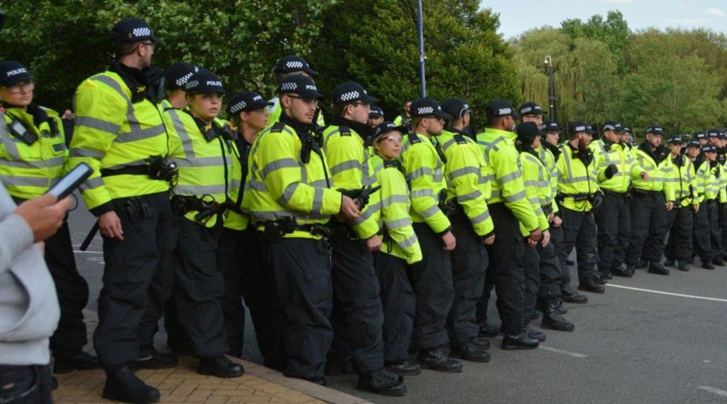 A row of dozens of police officers, all in hi-viz jackets, standing across a street. There are trees visible in the background