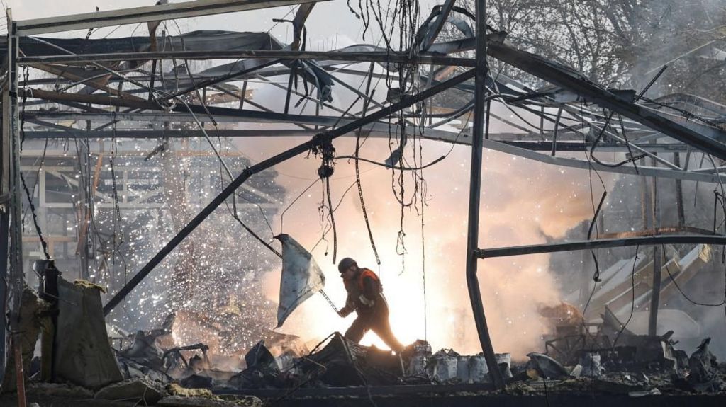 A rescue worker in the middle of rubble at a commercial building where only mangled metal frames remain; fire and smoke are shown in the background in the huge void betwen buldings