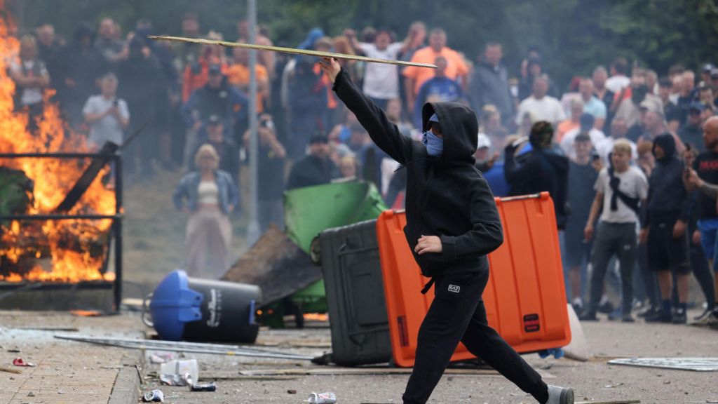 A man in a black hoodie with his face covered by sunglasses and a balaclava throws a stick, while in the background are overturned bins, a fire and a large crowd of people