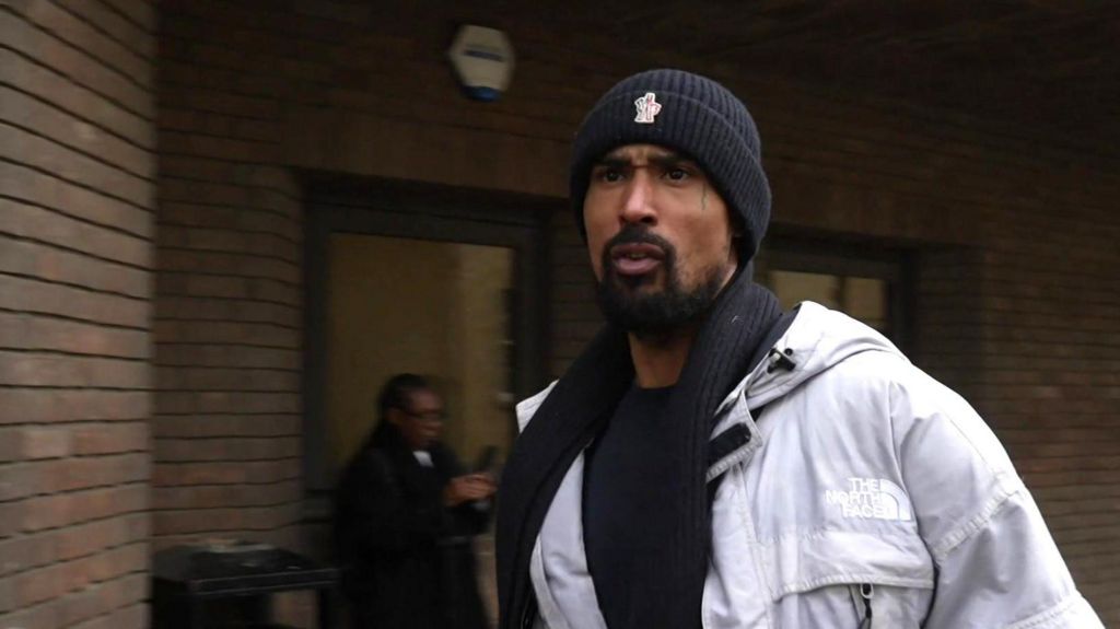 Ashley Warren walks outside the bricked frontage of Chelmsford Crown Court, wearing a white coat and dark-coloured beanie hat. He is mouthing something towards people behind the camera.