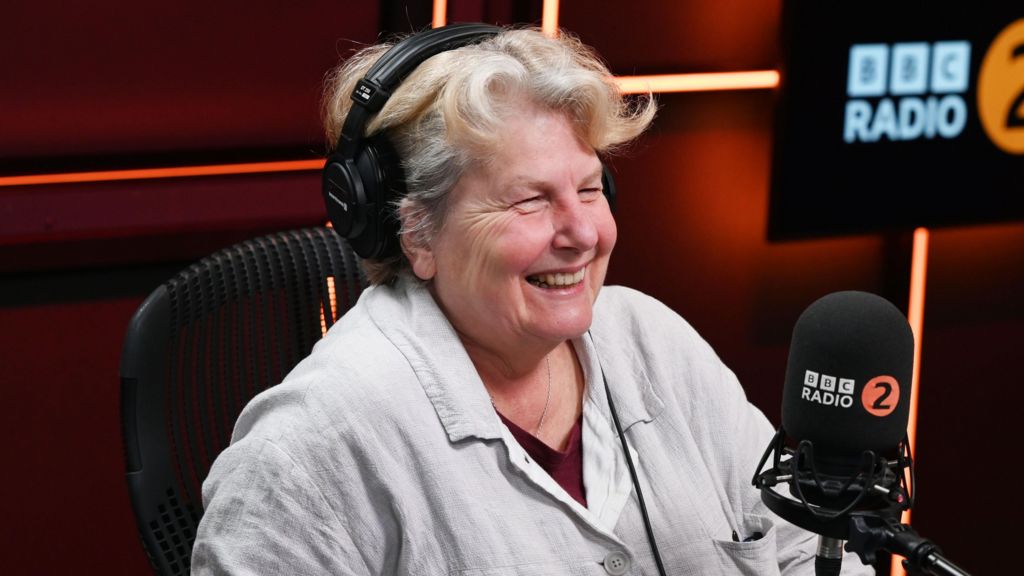 Sandi Toksvig is sitting an a BBC Radio 2 studio. In front of her is a black branded microphone and she is wearing headphones. The room is cast in dark orange lights. She is wearing a white shirt, red top and silver necklace. Toksvig  has short white and blonde hair and is smiling at someone off camera. 