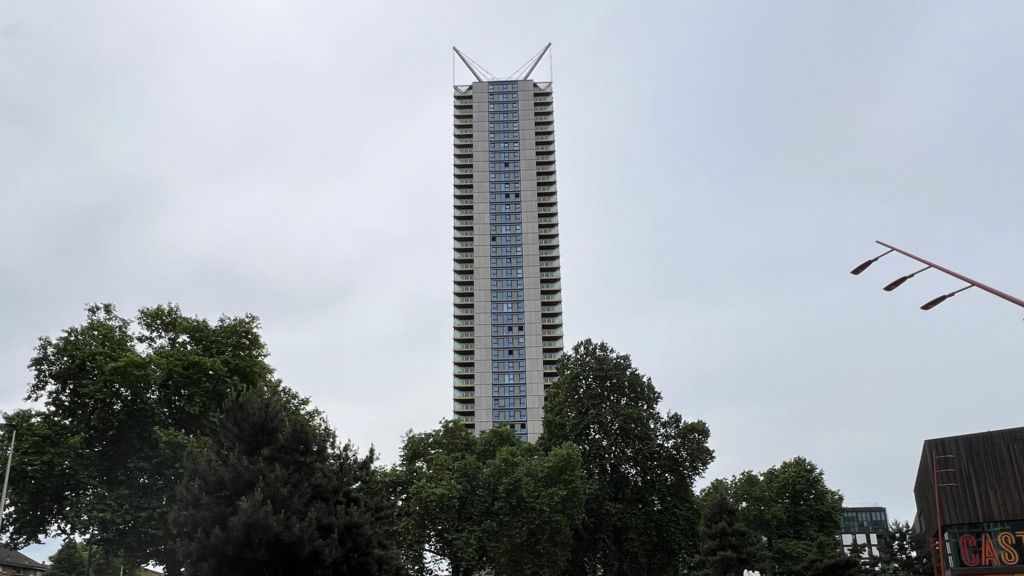 A tall, slender residential high-rise with distinctive V-shaped spires at the top, rising high above a line of green trees under an overcast sky.