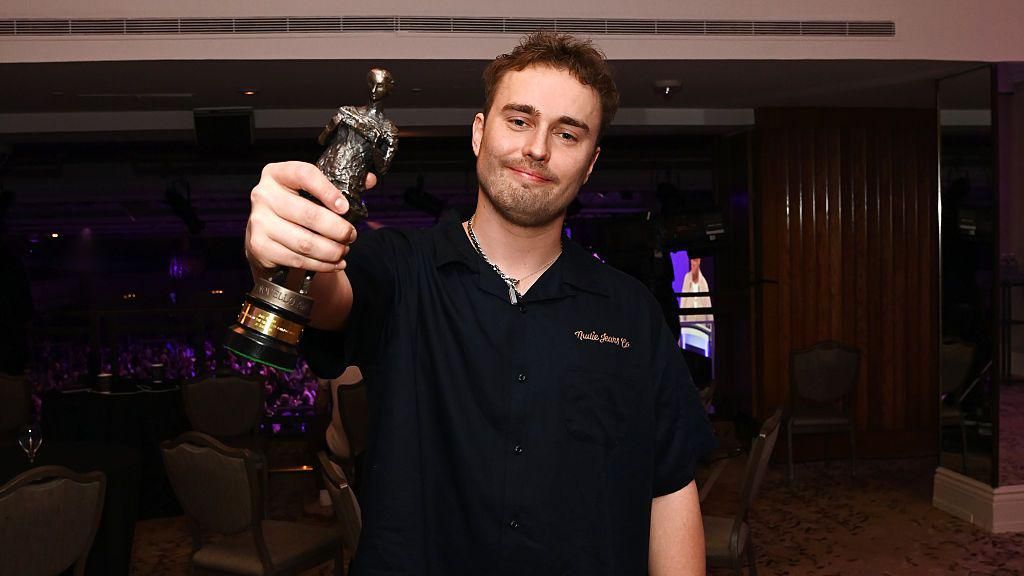 Sam Fender wears a black shirt and holds a statuette to the camera as he smiles. In the background is the ceremony hall