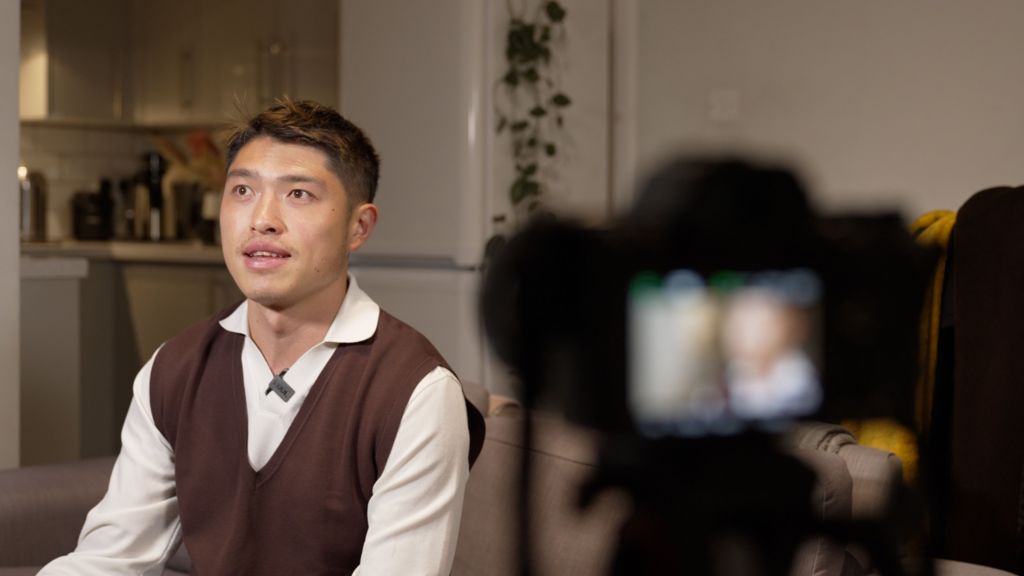 Andy Hu wears a white shirt and brown tank top. He is looking off to his right while a photographer frames him on a camera prior to filming. 