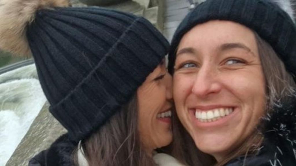 Amie Gray, a woman with blue eyes, brown hair and wearing a black woollen hat, smiles with her teeth. Her wife, Sian Gray, is also wearing a black woollen hat, and is nuzzled into Amie's cheek while smiling.
