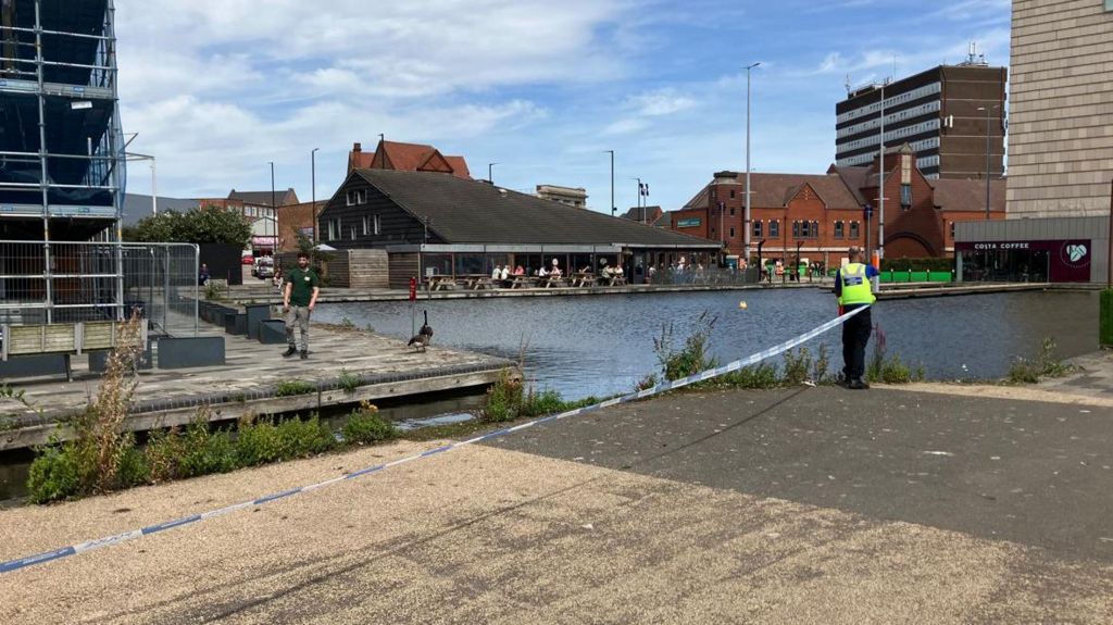 A photo of the canal at Walsall where a man in a high vis vest can be seen unravelling police cordon tape. Another man is walking on the opposite side of the canal. In the distance people sit outside a building (possibly a cafe or pub) and there are more buildings further in the distance