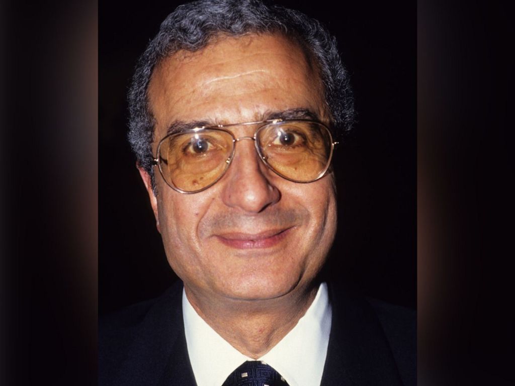 A photo of Ali Fayed with a sepia tone, taken several decades ago. He is smiling and wearing large tinted glasses. Behind is a treated image in green of Park Lane which takes in some of the greenery of Hyde Park.