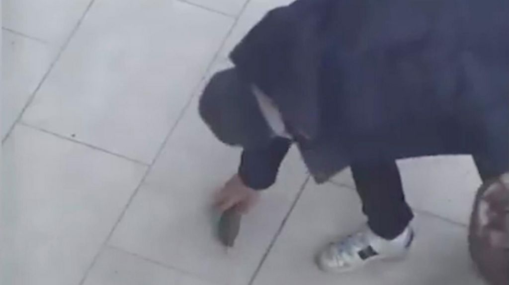 CCTV still of a man crouching on a tiled pavement as he places a small cylindrical object on the ground. He is wearing a dark coat and white trainers.