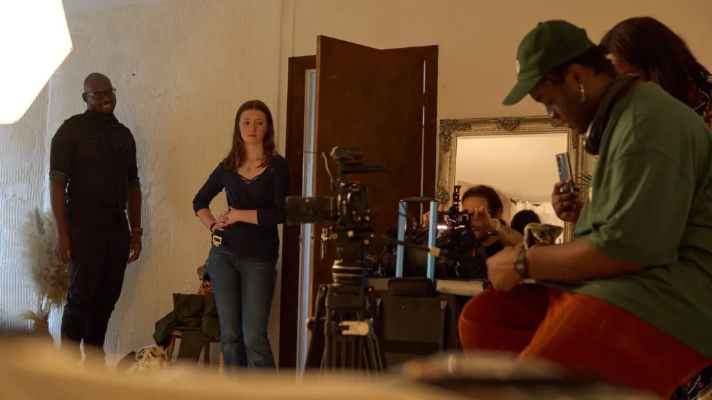 The crew on set of Rice at Home. There are multiple people in a room with a brown wooden door. A black man to the right of the photo, in a green shirt is controlling a camera. Various people are standing around, concentrating on a focal point off screen.
