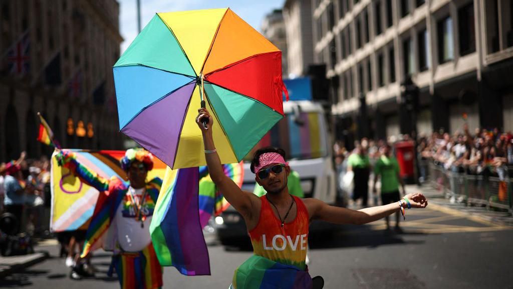 A person in a multi-coloured vest holding a rainbow umbrella dances in the parade