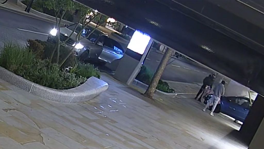 A car can be seen smashed into the front of a shop, with men surrounding it. The image is taken from CCTV footage which is taken on a slant.